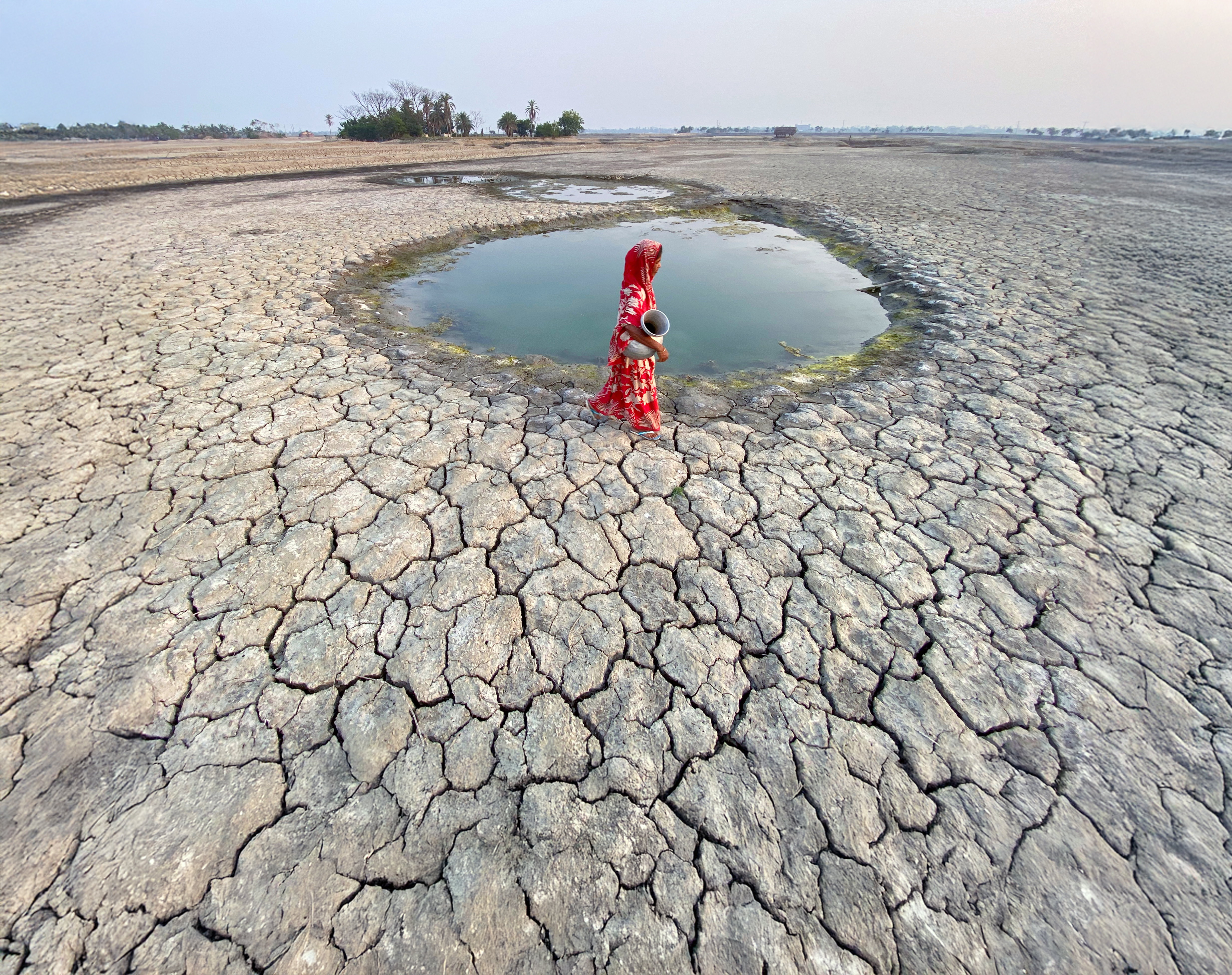 Woman carrying a water pot walks across dry, cracked earth.
