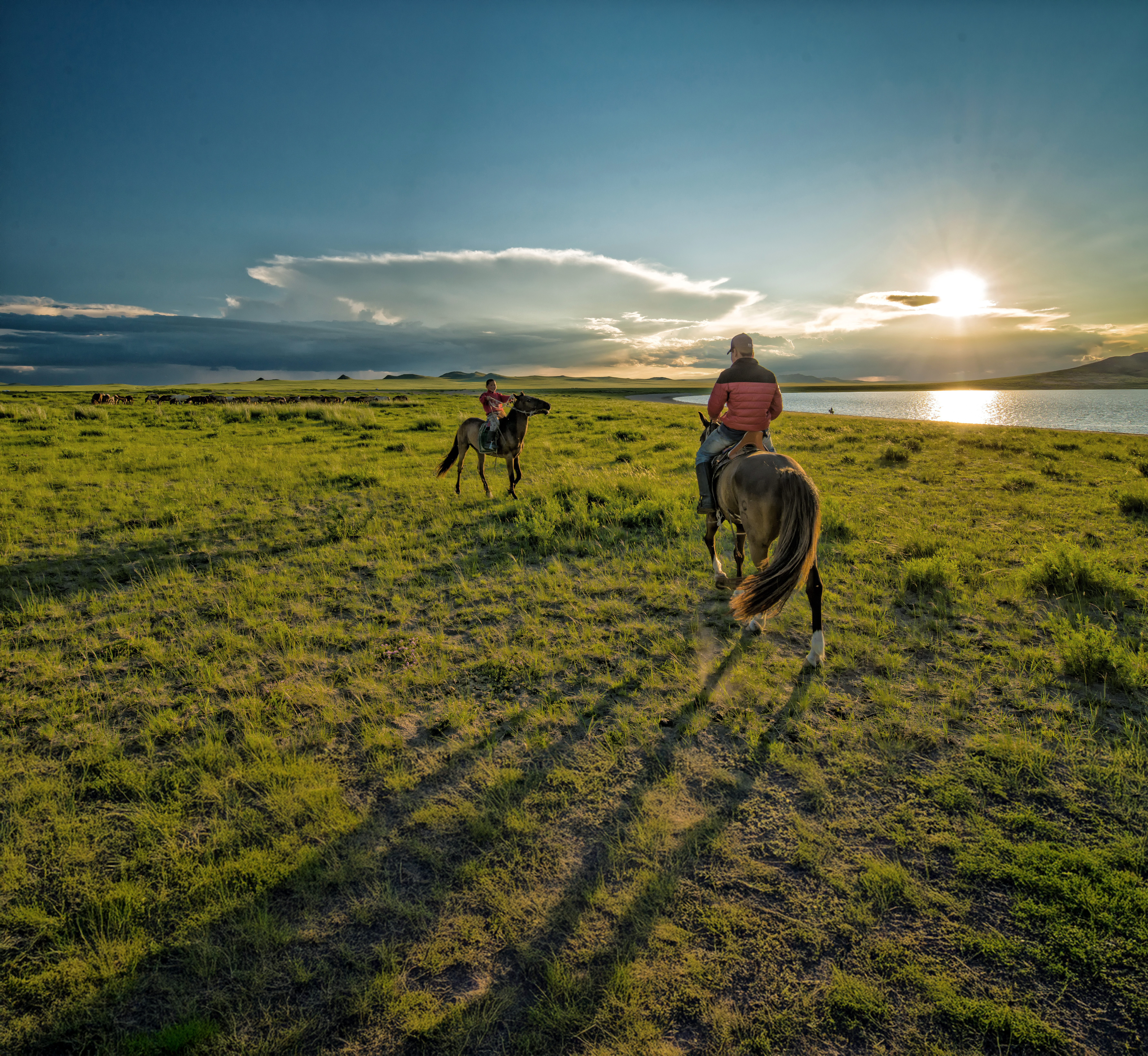 Two horseback riders on a grassy field near a lake.