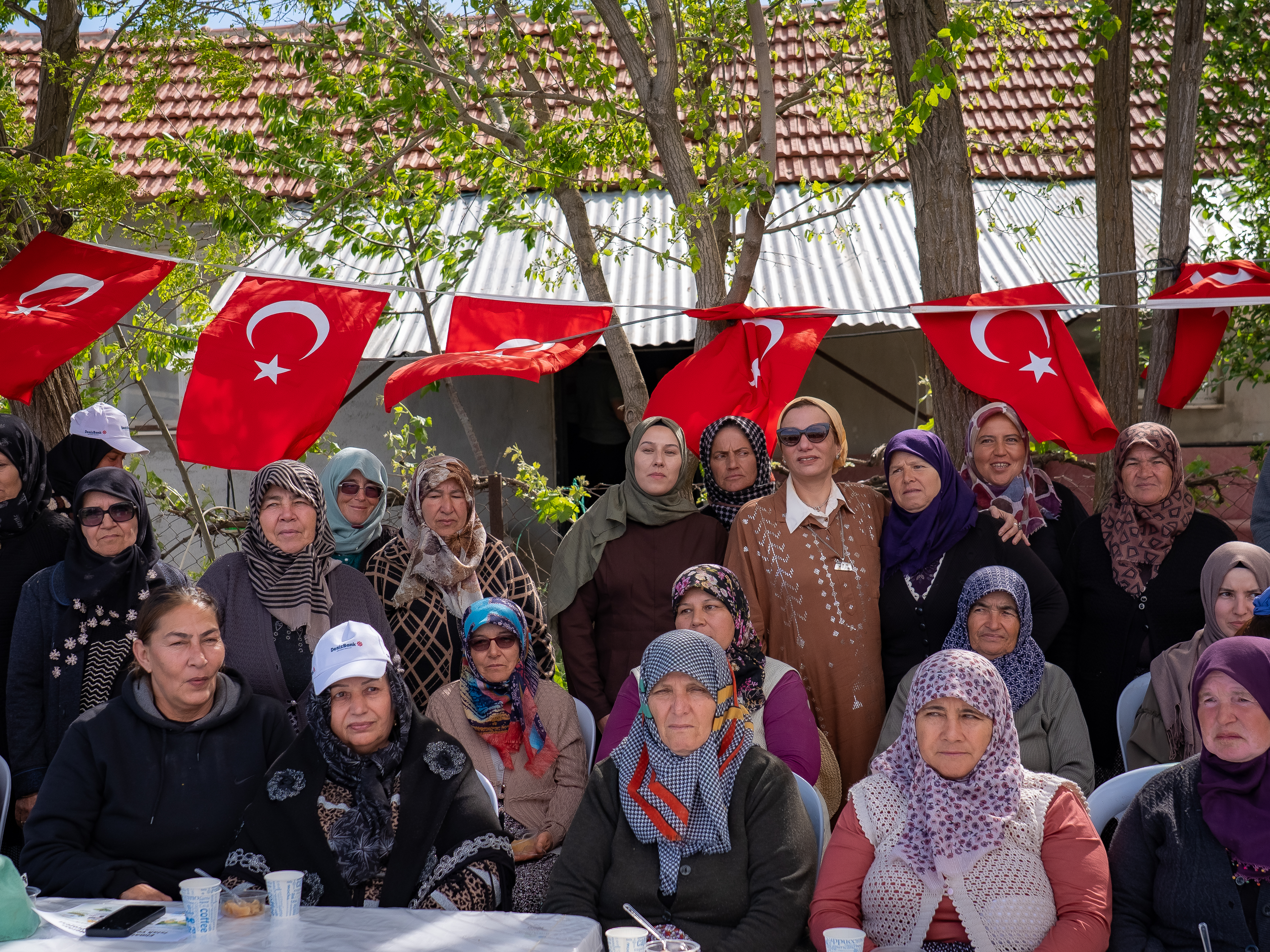 A group of women, many wearing headscarves, stand together outdoors beneath a string of vibrant Turkish flags.
